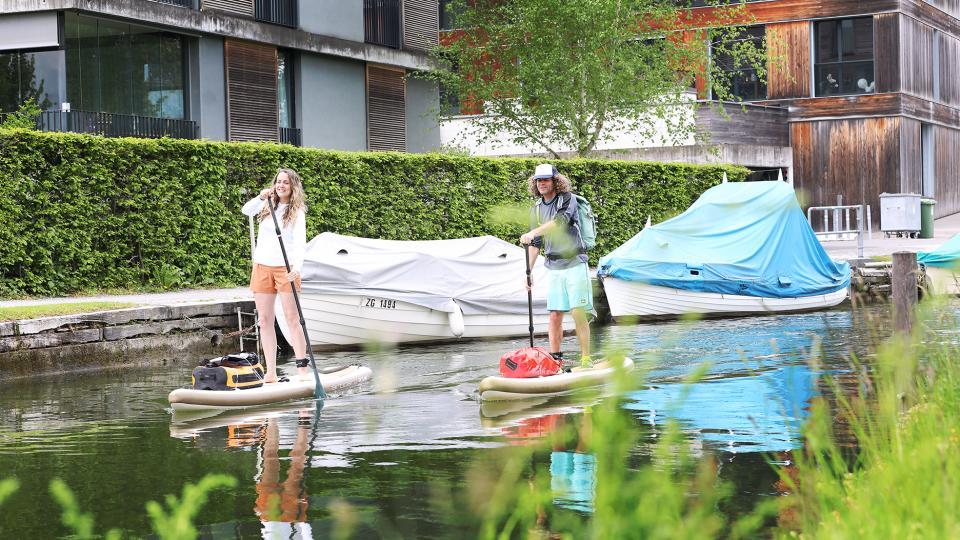 Zwei Paddelnde sind auf dem Lorzenkanal in Unterägeri unterwegs und paddeln vor Häusern und kleinen Booten durch.